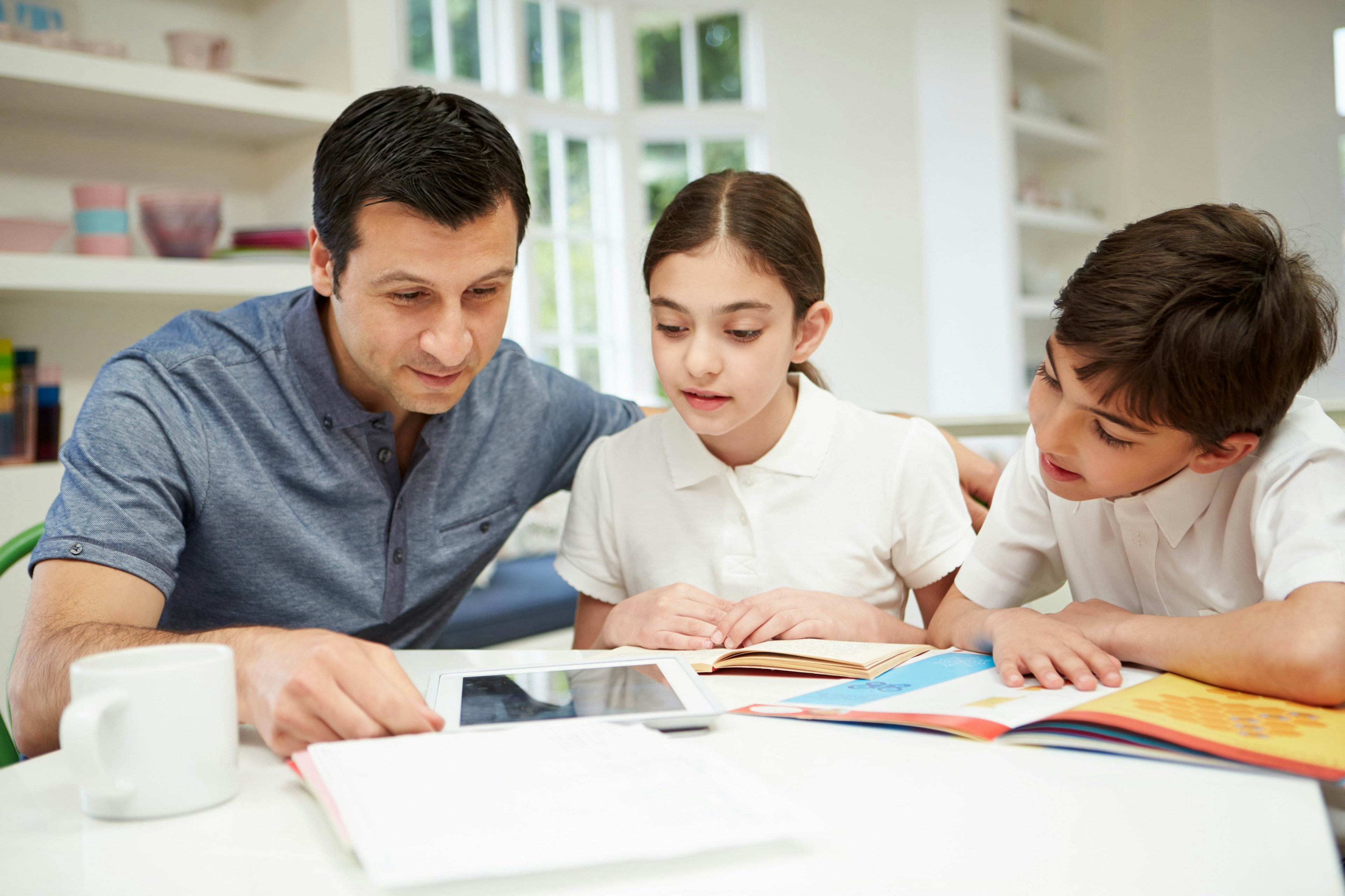 A man and two children look at books and a tablet on the table.