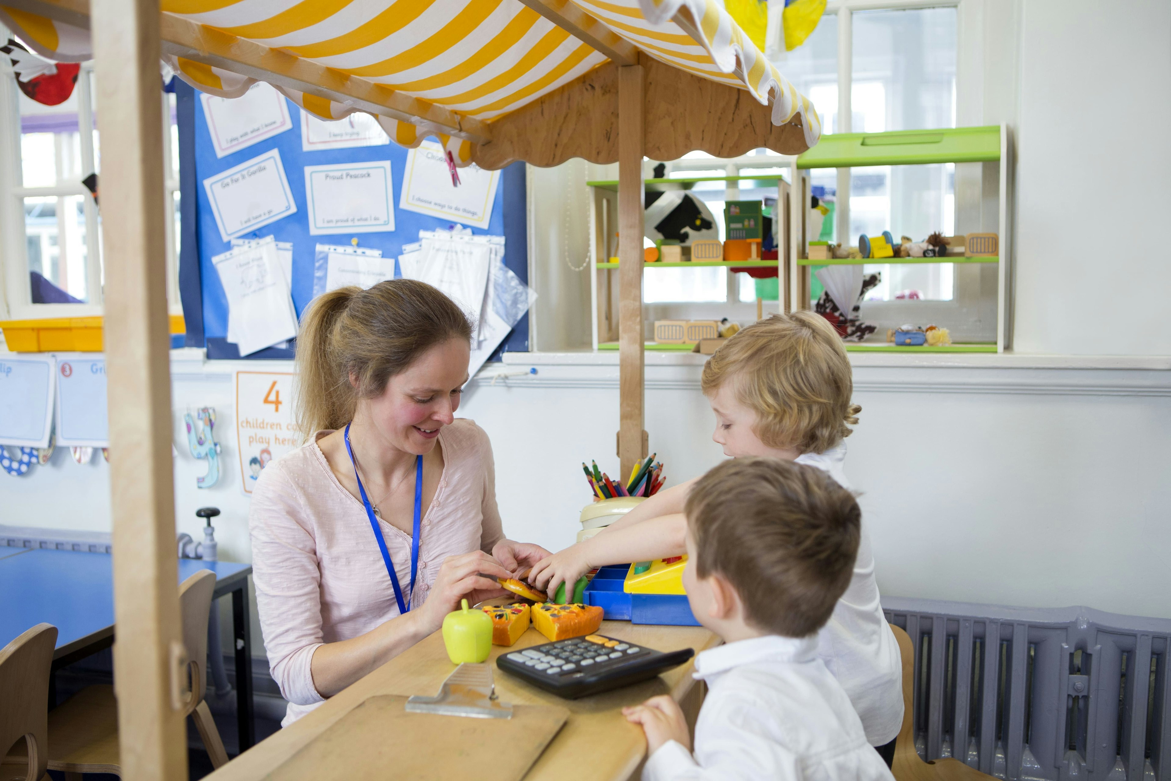 A teacher and two young children play in a role play shop with toy till.